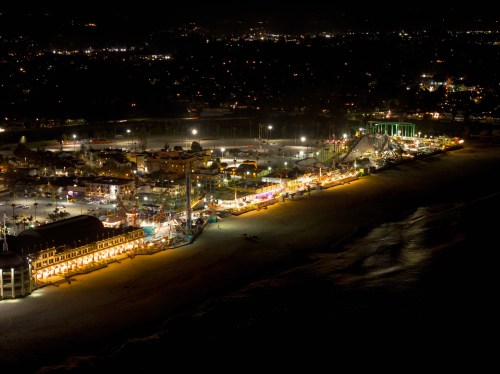 Aerial view of a vibrant beach amusement park at night, illuminated by colorful lights along the shoreline.