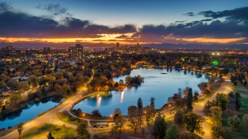 Aerial view of a serene lake surrounded by trees and city skyline at sunset, with colorful clouds and lights.