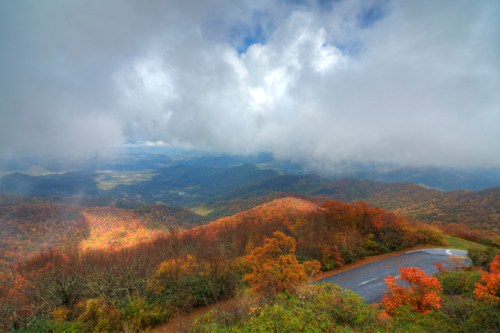 A panoramic view of colorful autumn foliage on mountains under a cloudy sky, with a winding road in the foreground.