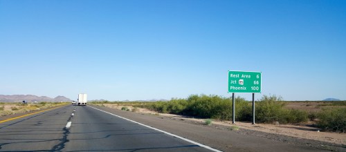 Highway scene with a green sign indicating a rest area in 6 miles, junction 66, and Phoenix 100 miles ahead.
