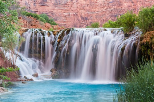 A serene waterfall cascades into a turquoise pool, surrounded by lush greenery and red rock formations.