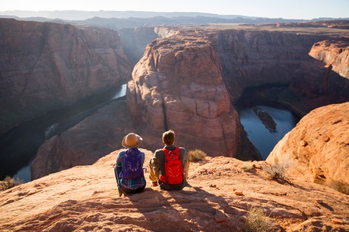 Two hikers sit on a rocky ledge, overlooking a winding river and canyon at sunset.