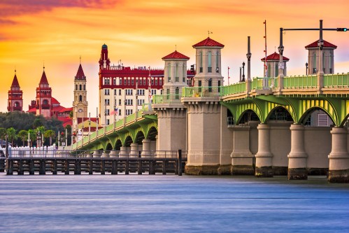 A scenic view of a green bridge at sunset, with historic buildings and a calm waterway in the foreground.