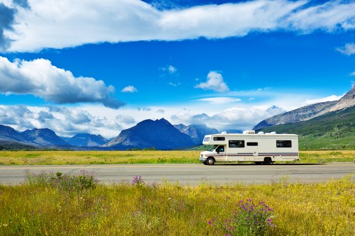 A white RV parked on a scenic road with mountains and blue skies in the background, surrounded by green fields and wildflowers.