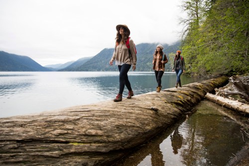 Three women walk along a large log by a serene lake, surrounded by mountains and trees under a cloudy sky.