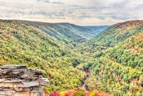 A panoramic view of a lush green valley with autumn foliage, framed by rocky cliffs under a cloudy sky.