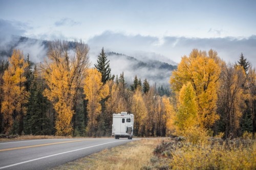 A camper van drives along a road lined with vibrant autumn trees and misty mountains in the background.