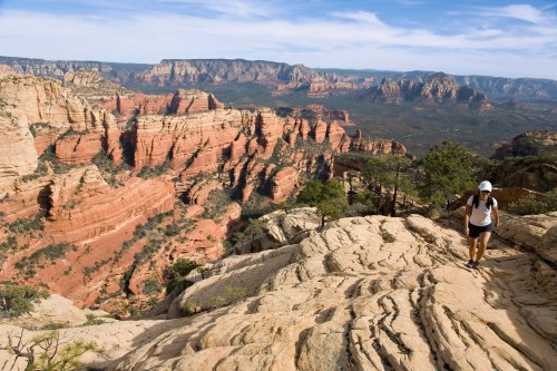 A hiker walks along rocky terrain with red rock formations and mountains in the background under a blue sky.