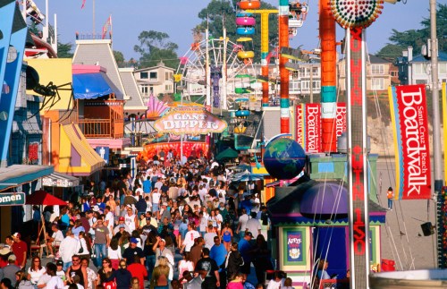 A bustling boardwalk scene filled with people, colorful shops, and amusement rides under a clear blue sky.