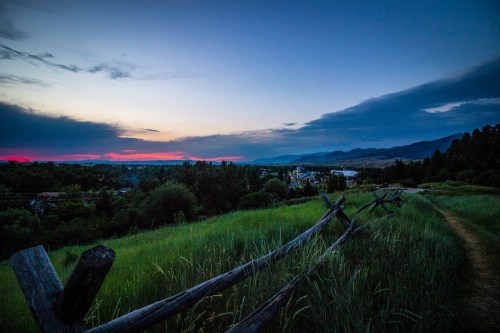A serene landscape at dusk, featuring a grassy field, a wooden fence, and distant mountains under a colorful sky.