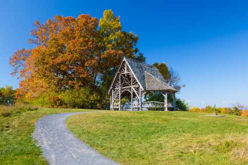 A wooden gazebo on a grassy hill, surrounded by colorful autumn trees under a clear blue sky.