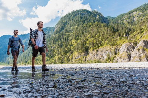 Two men hike along a rocky riverbank, surrounded by lush green mountains and a bright blue sky.