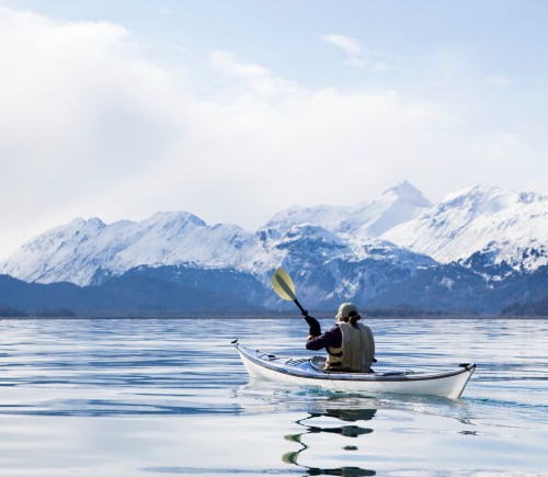 A person kayaking on calm water with snow-capped mountains in the background under a clear sky.