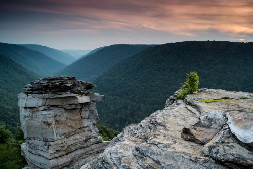 A rocky cliff overlooks a lush green valley under a cloudy sky at sunset.