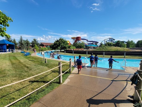A sunny water park scene with a lazy river, children playing, and water slides in the background. Green grass and blue sky.