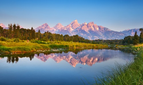 Grand Teton reflected in the calm waters of the Snake River at Schwabacher’s Landing in Wyoming, surrounded by trees and mountain scenery.