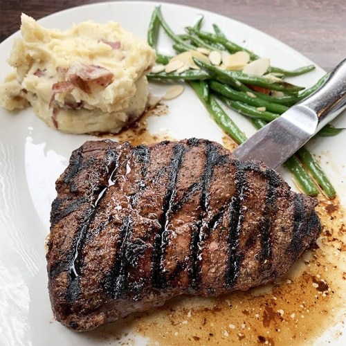 A grilled steak with a knife, served alongside mashed potatoes and green beans with almonds on a white plate.