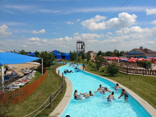 A sunny water park scene with people floating in a lazy river, surrounded by greenery and colorful umbrellas.