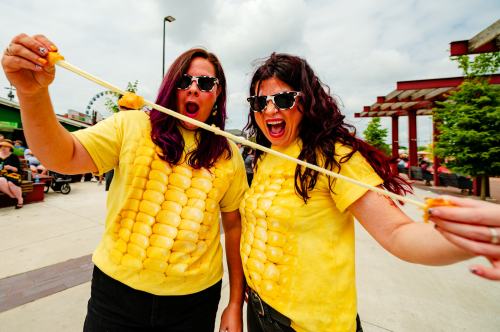 Two women in corn-themed shirts excitedly pose, holding a long piece of cheese, with a fairground in the background.