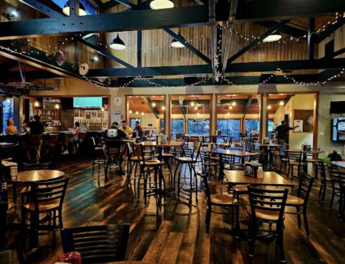 A spacious, warmly lit restaurant interior with wooden floors, tables, and a bar area, featuring large windows and string lights.