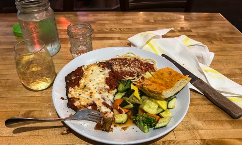 A plate with baked pasta, marinara sauce, zucchini, broccoli, and garlic bread, with a glass of water nearby.