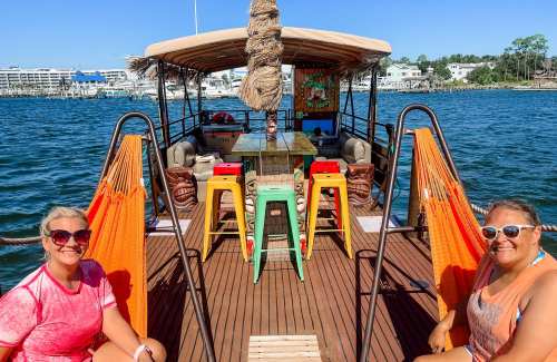 Two women relax on a colorful boat with hammocks, surrounded by water and a sunny sky.