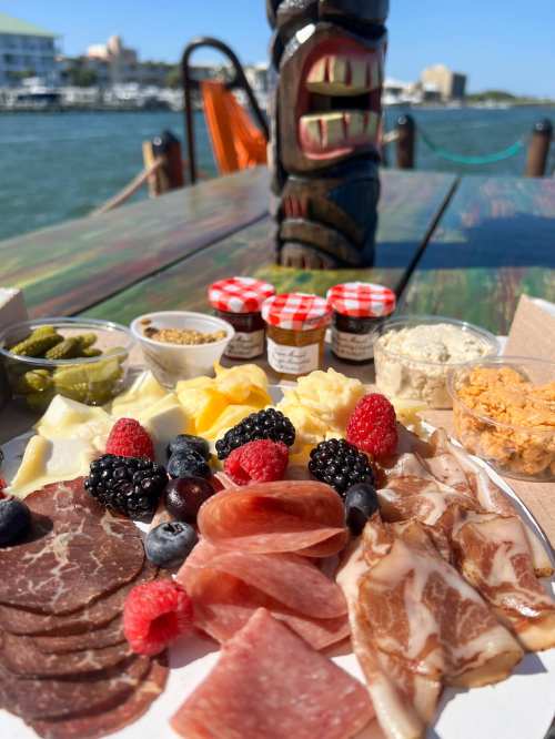 A colorful charcuterie board with meats, cheeses, fruits, and condiments, set against a waterfront backdrop.
