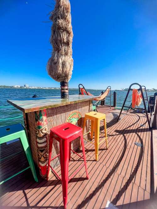 A colorful bar with stools on a deck by the water, featuring a tiki-style design and clear blue skies.