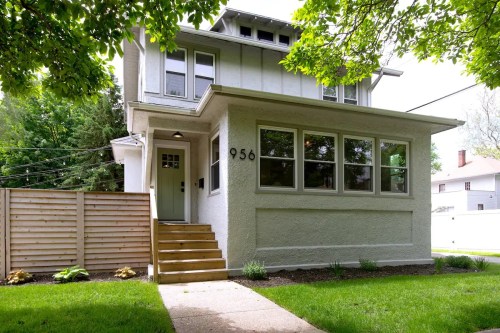 A two-story white house with large windows, a wooden staircase, and a fenced yard, surrounded by greenery.