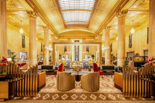 Luxurious hotel lobby with golden decor, tall columns, elegant seating, and large windows allowing natural light.