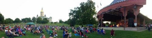 A large crowd enjoys an outdoor concert at a park, with a historic building visible in the background.