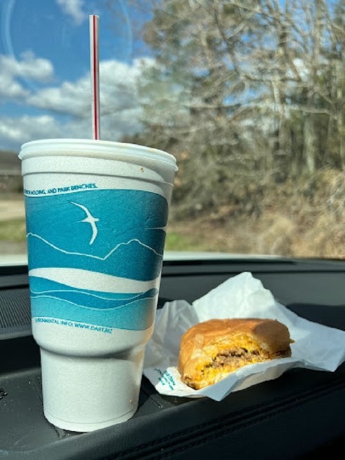 A drink in a paper cup and a wrapped sandwich on a car dashboard, with trees and a blue sky in the background.