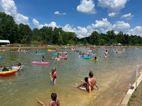 A crowded lake scene with people swimming and floating on colorful inflatables under a sunny sky with fluffy clouds.