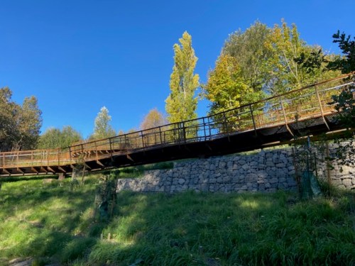 A rust-colored bridge spans a grassy area, surrounded by trees under a clear blue sky.