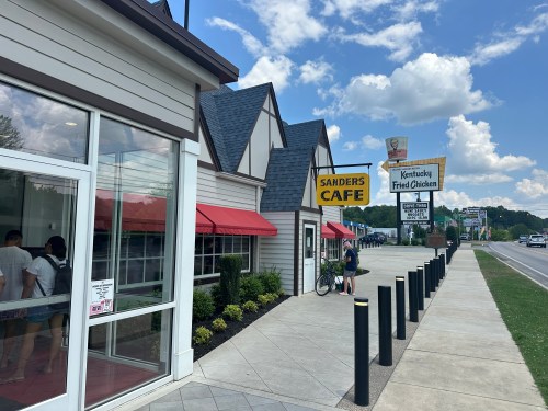 Exterior of Sanders Cafe with a sign for Kentucky Fried Chicken, featuring a sunny day and a sidewalk.