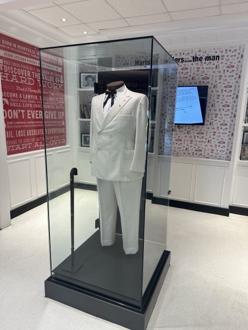 A white suit displayed in a glass case, with a black bow tie, in a museum setting with red and white wall graphics.