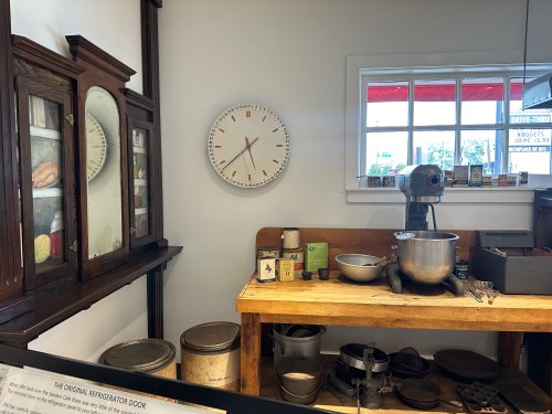 A vintage kitchen scene featuring a clock, mixer, and various cooking utensils on a wooden table.