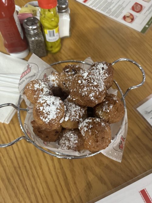 A basket of golden-brown apple fritters dusted with powdered sugar, placed on a wooden table.