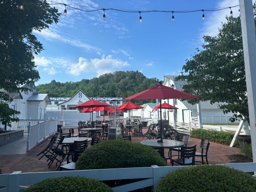 Outdoor dining area with red umbrellas, tables, and a scenic backdrop of trees and blue sky.