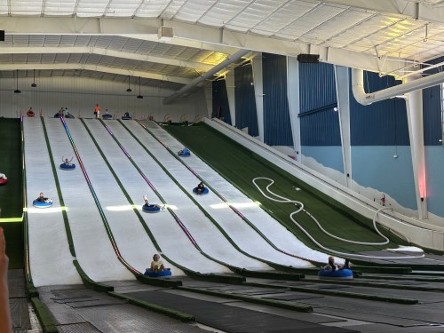 Indoor snow tubing hill with multiple lanes and people sliding down on inflatable tubes. Brightly lit and spacious.