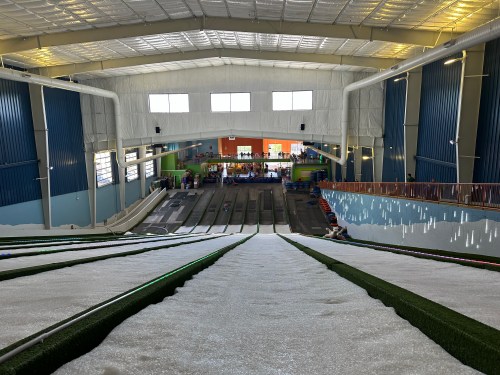 View from the top of an indoor snow tubing slope, with multiple lanes and colorful walls in the background.