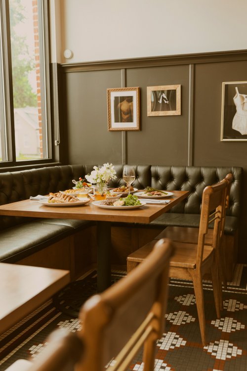 A cozy restaurant table set with various dishes, flowers, and wine, surrounded by elegant decor and natural light.