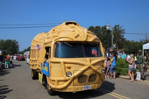 A whimsical peanut-shaped vehicle drives through a parade, surrounded by onlookers and festive decorations.