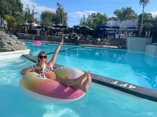 A person relaxes on a float in a pool, smiling and giving a thumbs up, with palm trees and umbrellas in the background.