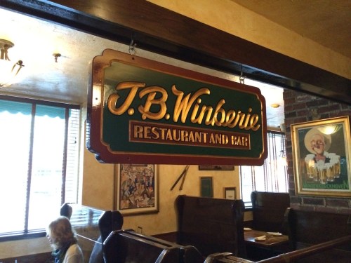 Sign for J.B. Winberie Restaurant and Bar, hanging in a cozy interior with wooden booths and warm lighting.