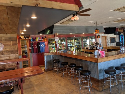 Interior of a rustic bar with a wooden counter, stools, and shelves filled with drinks. Casual and inviting atmosphere.