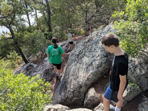 Three people hiking on rocky terrain surrounded by trees and greenery.