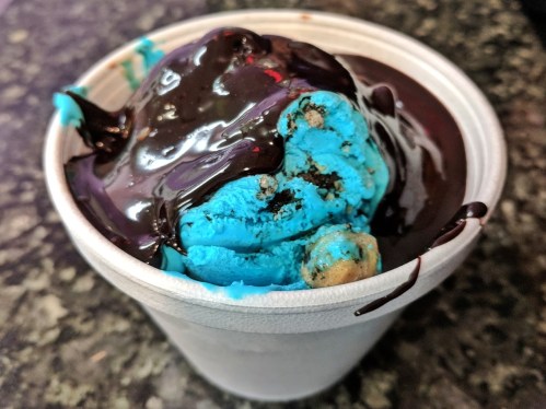 A cup of bright blue ice cream topped with chocolate sauce and cookie pieces.