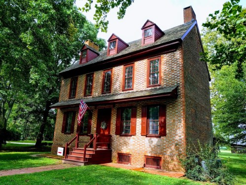 A historic brick house with red shutters and an American flag, surrounded by green trees and a grassy lawn.