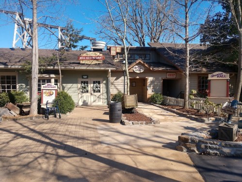 A rustic restaurant building with a sign, surrounded by trees and a paved pathway on a sunny day.